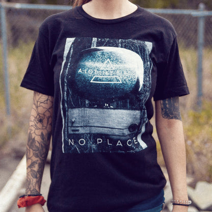 Black short sleeve shirt with an image with an old TV in the center. There is static on the TV and text in it that says A LOT LIKE BIRDS. Below the TV there is text that says NO PLACE. An individual is modeling the shirt and standing in front of a fence with grass in the background.