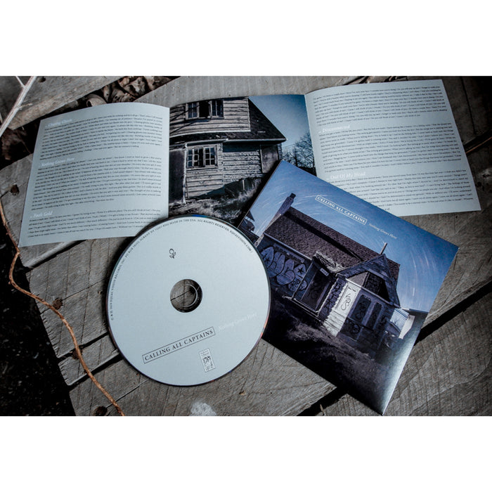 Square CD with an image of a house with graffiti on the back. The sun is casting shadows over the house. There is white text in the sky that says CALLING ALL CAPTAINS. There is a white CD laying on top of it and both are on top of a wooden surface.