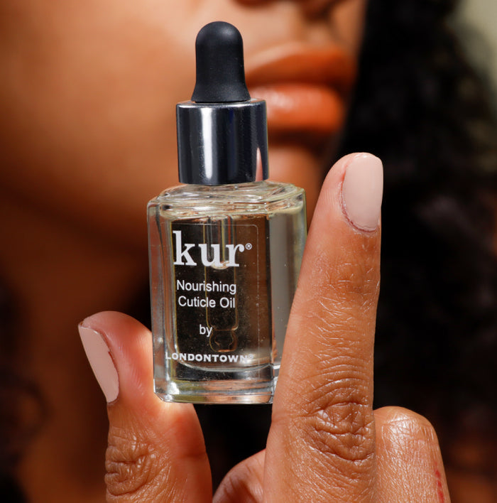 Close-up of a person holding a bottle of Kur Nourishing Cuticle Oil by Londontown. Their nails are polished in a soft pink shade.