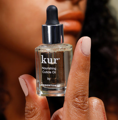 Close-up of a person holding a bottle of Kur Nourishing Cuticle Oil by Londontown. Their nails are polished in a soft pink shade.