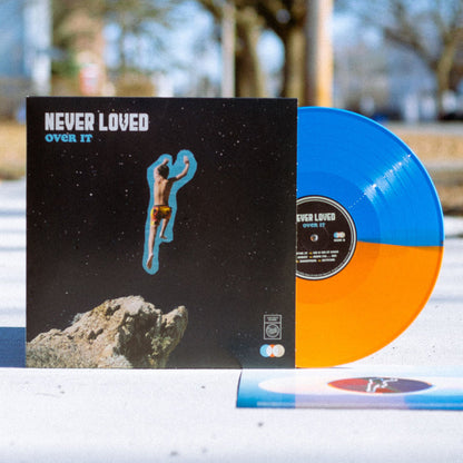 Vinyl jacket with NEVER LOVED written in the top left corner in white font. Below that there is blue text that says OVER IT. The album artwork of a boy jumping off of a cliff, but in a starry space sky. Peeking out of the jacket is a half blue and half orange vinyl. Both are standing up on top of a sidewalk outside with trees in the background.