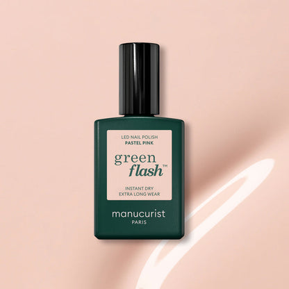 A bottle of Manucurist Pastel Pink gel polish with beige undertones, dark green cap and label, sits on a light pink surface beside a curved white reflection.