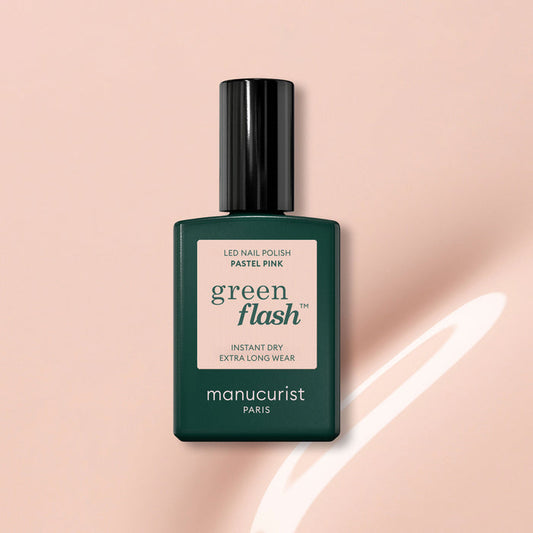 A bottle of Manucurist Pastel Pink gel polish with beige undertones, dark green cap and label, sits on a light pink surface beside a curved white reflection.