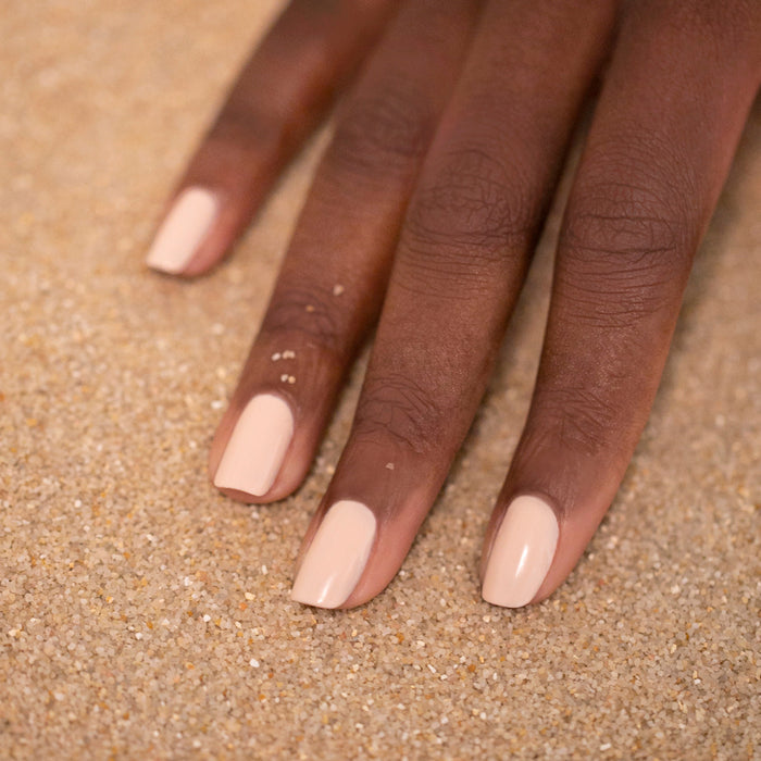 A hand with medium-dark skin tone rests on sand, featuring neatly manicured nails painted in Manucurist's Pastel Pink—a glossy nude shade with subtle beige undertones.