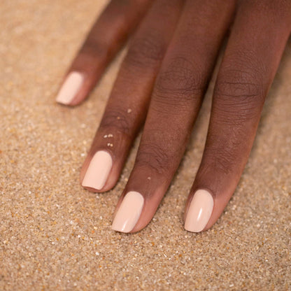 A hand with medium-dark skin tone rests on sand, featuring neatly manicured nails painted in Manucurist's Pastel Pink—a glossy nude shade with subtle beige undertones.
