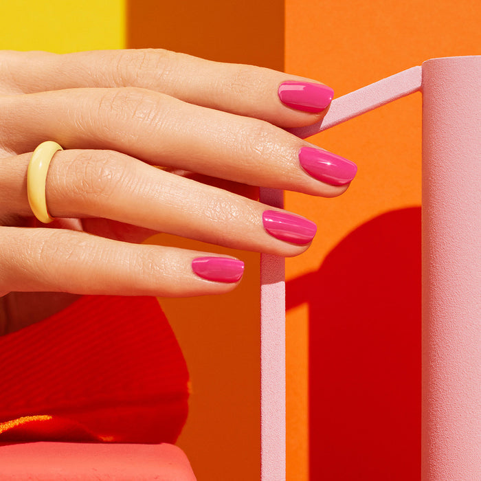 A hand with glossy pink Pétula Green Flash gel polish by Manucurist and a yellow ring rests on pink geometric shapes, set against a vibrant orange-red background—the perfect summer nail look.
