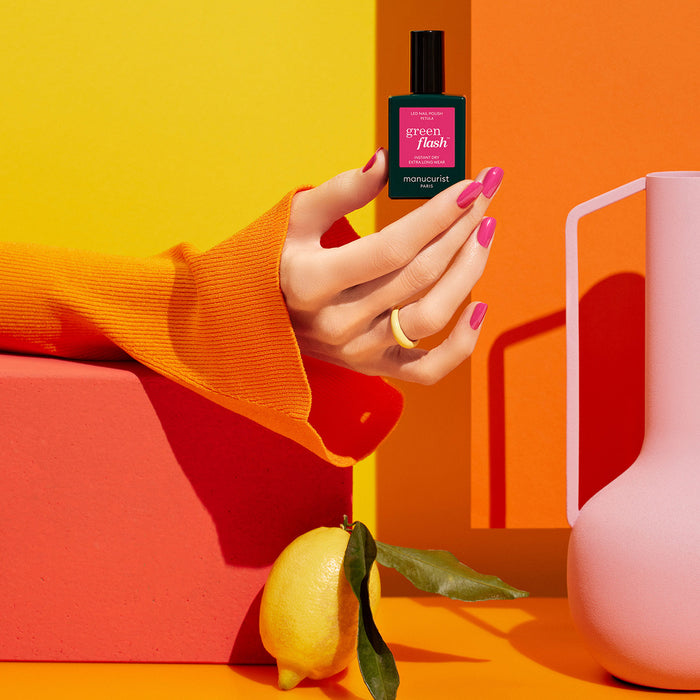 A hand with Pétula by Manucurist Green Flash gel polish holds a green nail polish bottle against an orange-yellow background, with a lemon and pink pitcher nearby. The hand, wearing an orange sleeve and white ring, rests on a red block.