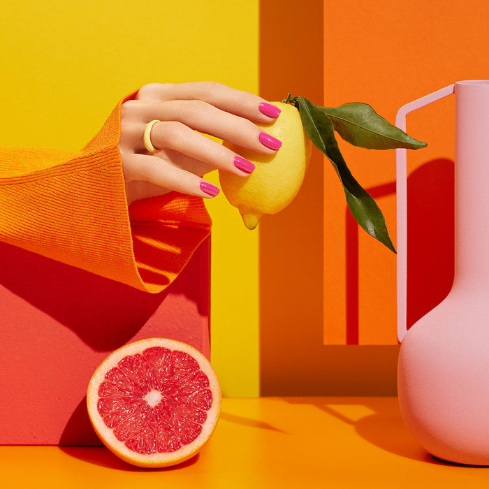A hand with Manucurist’s Pétula Green Flash pink gel polish and an orange sleeve holds a lemon. A pink grapefruit half and a pink vase rest on a bold yellow and orange geometric background.