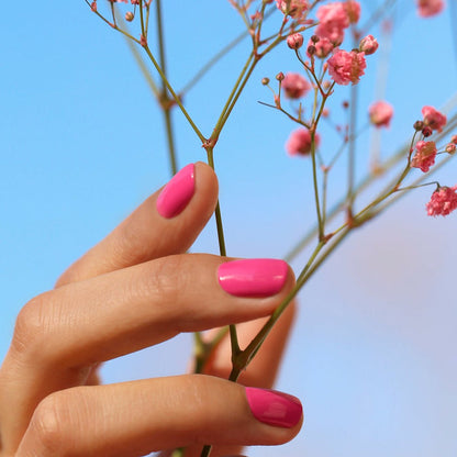 A hand with Manucurist's Pétula Green Flash gel polish—a bright pink shade with blue undertones—holds delicate pink flowers against a clear blue sky.