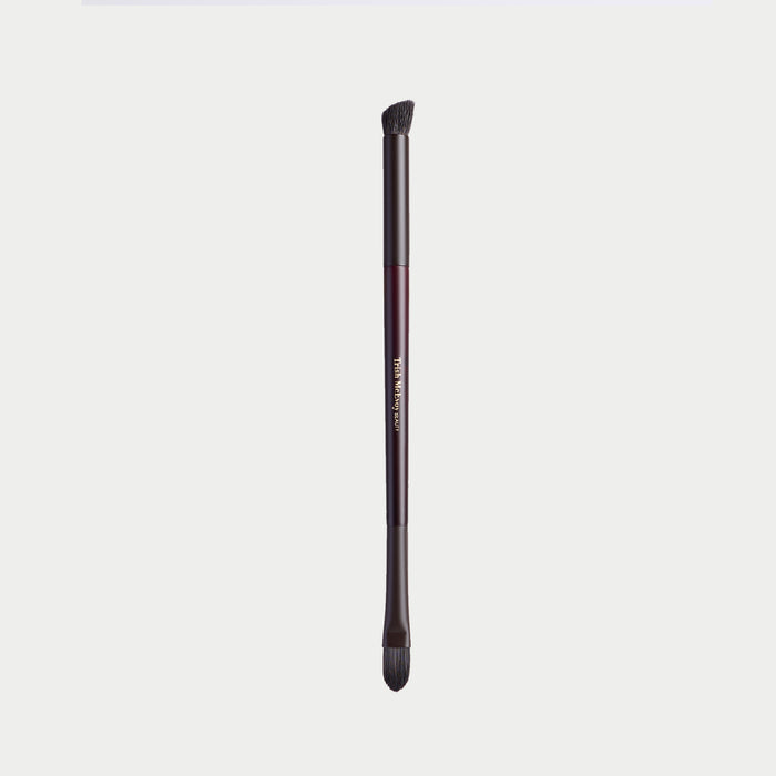 A single makeup brush with a dark metallic handle and black bristles, shown vertically against a light gray background. The handle features a metallic rose gold accent band with gold lettering that reads 'm.