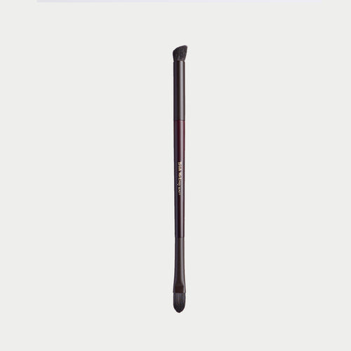 A single makeup brush with a dark metallic handle and black bristles, shown vertically against a light gray background. The handle features a metallic rose gold accent band with gold lettering that reads 'm.