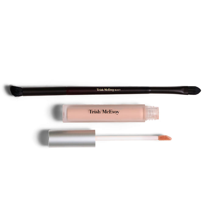 Trish McEvoy makeup set featuring a dual-ended applicator brush, a clear tube of liquid product in a nude pink shade, and its silver cap and doe-foot applicator wand, all displayed on a clean white background.