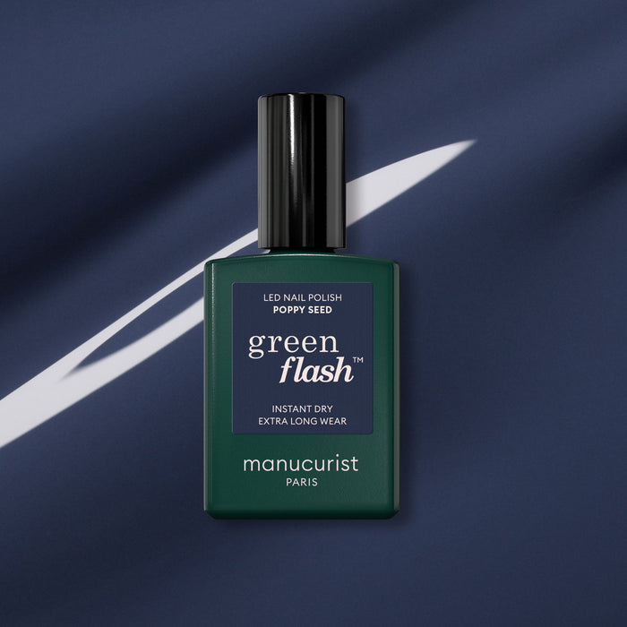 A dark green bottle of Manucurist Poppy Seed gel polish is shown against a dark blue fabric background with a white diagonal streak.