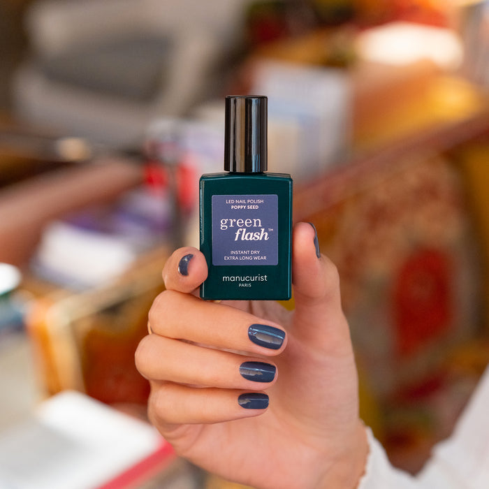 A hand with neatly manicured nails painted in the chic Poppy Seed shade holds a bottle of Manucurist's Green Flash LED nail polish. The background is softly blurred in warm tones.
