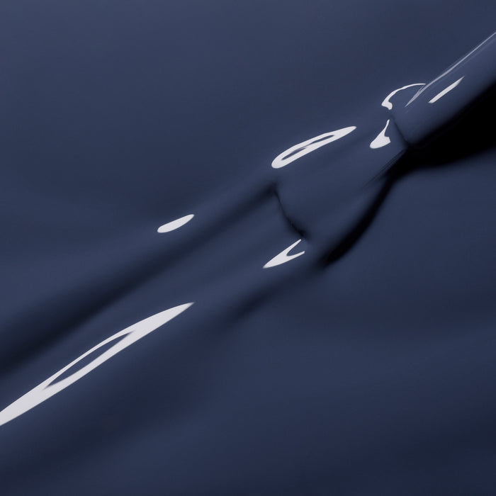 A close-up of Poppy Seed by Manucurist shows dark blue liquid with a smooth, glossy surface and reflective highlights, forming abstract shapes reminiscent of bluish grey nail polish.