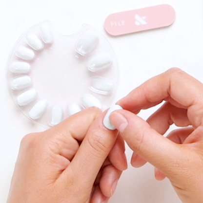 Hands holding a white press-on nail near a circular display with white false nails and a pink nail file on white background