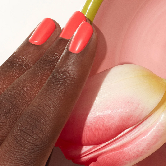 A hand with dark skin wears Manucurist's Pulp, a bright coral pink gel polish, resting gently on a pink and white tulip petal against a soft pink background.