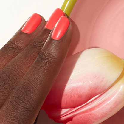 A hand with dark skin wears Manucurist's Pulp, a bright coral pink gel polish, resting gently on a pink and white tulip petal against a soft pink background.