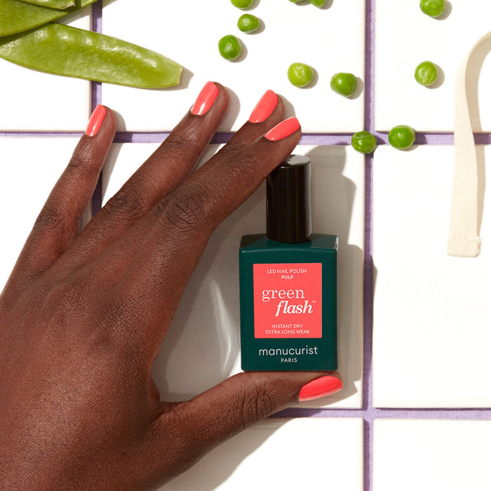 A hand with coral pink gel polish holds a Manucurist Pulp bottle on a white tiled surface, surrounded by green peas and snap pea pods.