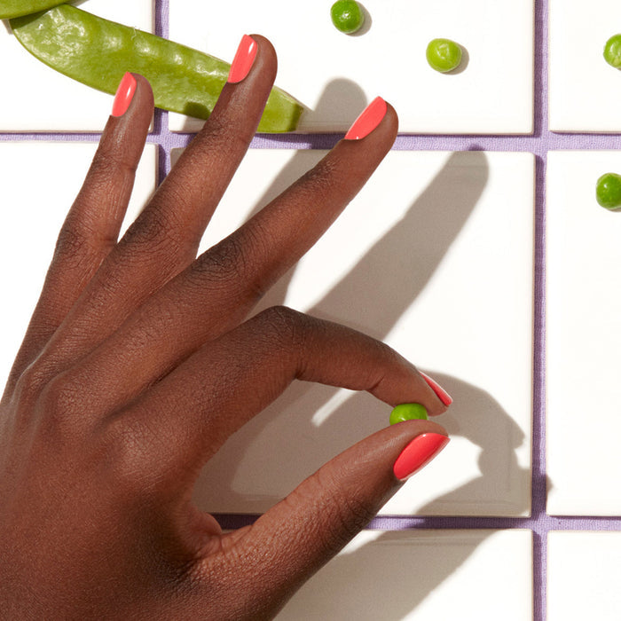 A hand with Manucurist's Pulp coral pink gel polish holds a single green pea above white tiles, surrounded by more peas and a pea pod.
