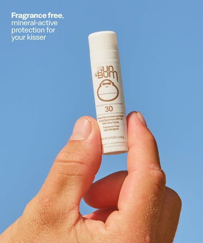 Hand holding a Sun Bum lip balm against a blue background. Fragrance free, mineral-active protection for your kisser. Fragrance free, mineral-active protection for your kisser