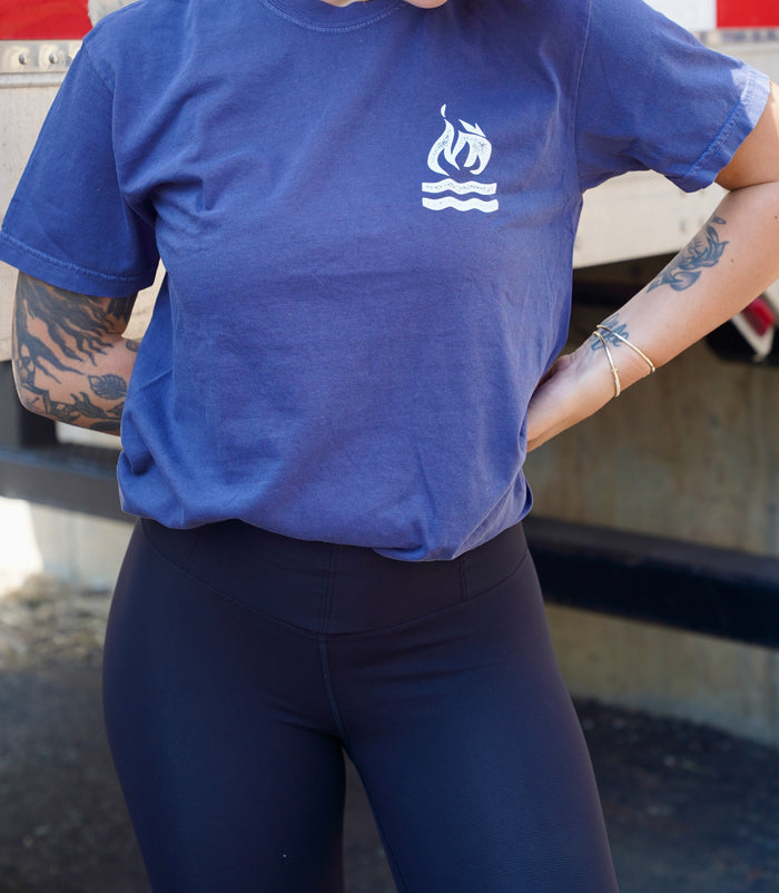 A person wearing a blue, garment-dyed Hot Water Music Comfort Colors T-Shirt with a white boat logo and black leggings stands outdoors, showing tattooed arms and silver bracelets. Their head is out of the frame.