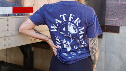 A tattooed person wears a navy Comfort Colors T-shirt by Hot Water Music, featuring a white rose with water drops and bold "WATER MUSIC" text. They stand outdoors near metal, resting one hand on their hip.