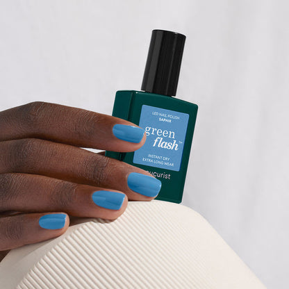 A hand with medium-dark skin tone displays neatly manicured nails painted in Saphir by Manucurist, holding a matching bottle of Manucurist Saphir gel polish against a light background.