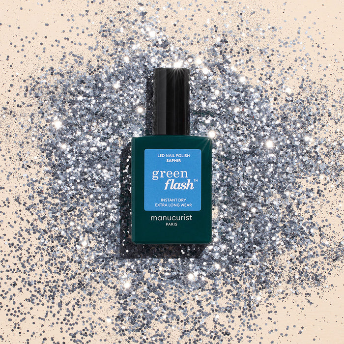 A teal bottle of Manucurist Saphir gel polish rests on a surface dusted with shiny silver glitter, creating a sparkling, glamorous look.