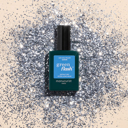 A teal bottle of Manucurist Saphir gel polish rests on a surface dusted with shiny silver glitter, creating a sparkling, glamorous look.