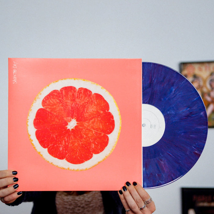 Bright orange colored vinyl jacket with an image of the inside of a grapefruit. In the top left corner there is vertical white text that says SAVES THE DAY. Peeking out of the jacket is a purple vinyl. An individual is holding the jacket and standing in front of a white wall.