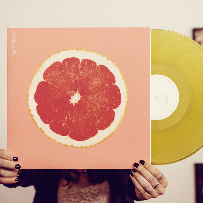 Bright orange colored vinyl jacket with an image of the inside of a grapefruit. In the top left corner there is vertical white text that says SAVES THE DAY. Peeking out of the jacket is a yellow vinyl. An individual is holding the jacket and standing in front of a white wall.