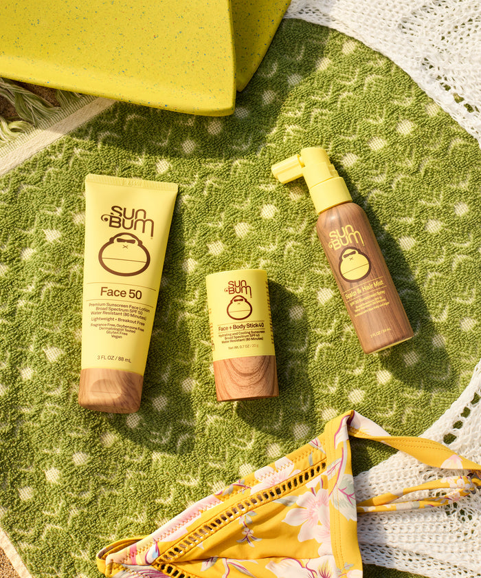 Scalp & Hair Mist SPF 30 spray bottle displayed with other Sun Bum sunscreen products on a green textured blanket alongside yellow beach accessories and white lace trim, showcasing a complete sun protection collection by Sun Bum