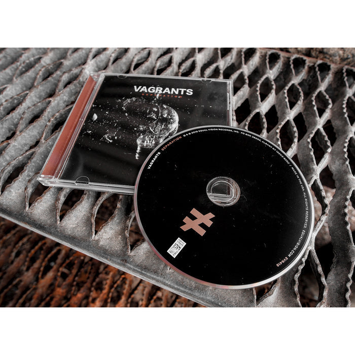 Square CD with VAGRANTS written in white font. Below that there is smaller font that says SEPARATION. The album art is a black and white image of a light bulb shattering. Laying on top of the cover is a black disc with a not equal to sign in the bottom. Both are laying on top of stairs.