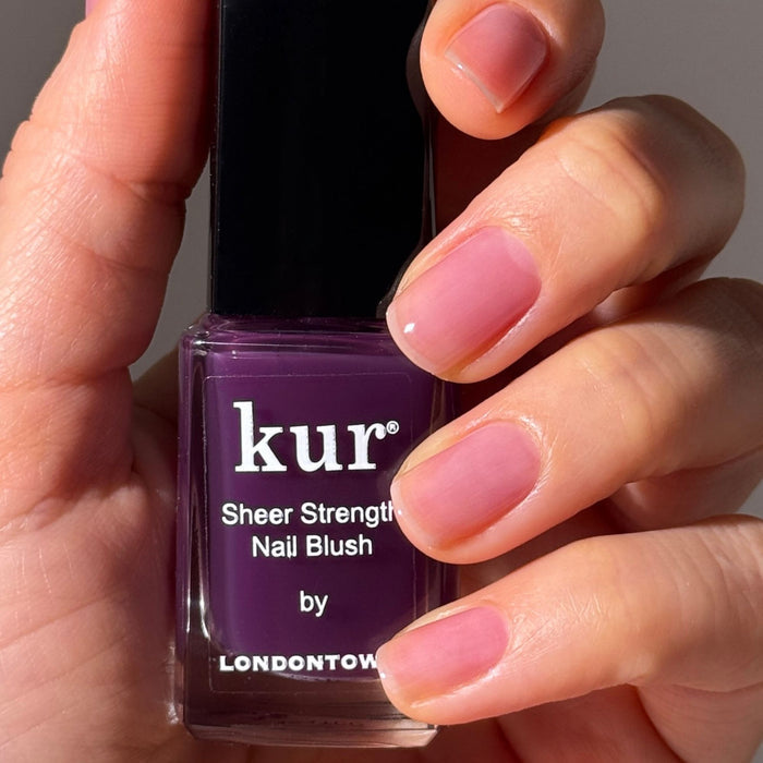 A hand holds a bottle of LONDONTOWN Sheer Strength Nail Blush - Concord, a 2-in-1 nail hardener and glow booster; nails are painted with sheer, natural pink polish.