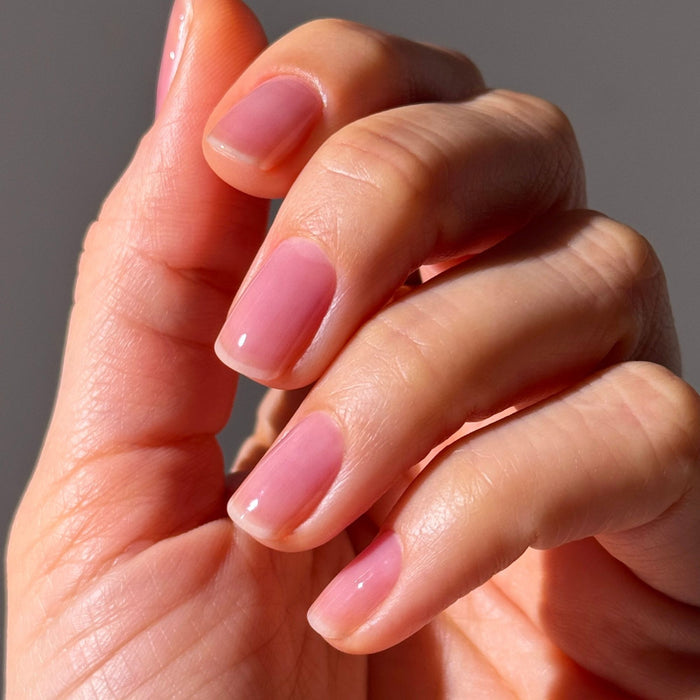 A close-up of a hand with short, manicured nails coated in LONDONTOWN Sheer Strength Nail Blush - Concord, a glossy light pink 2-in-1 nail hardener for extra strength and shine.