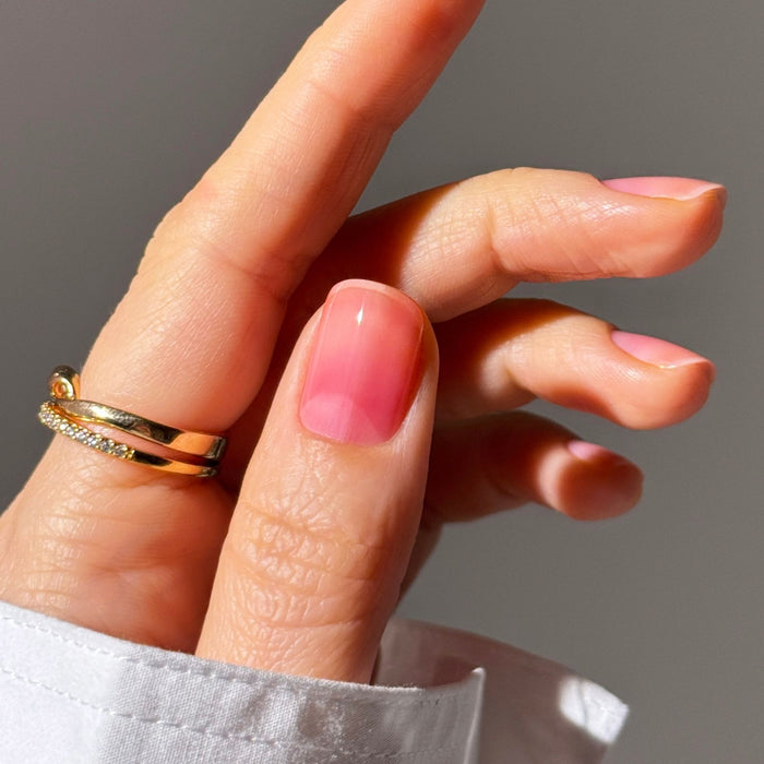 A hand with gold rings displays a manicured nail painted in LONDONTOWN Sheer Strength Nail Blush - Poppy, a glossy light pink polish with a glow boost, set against a neutral background.