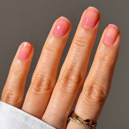 A hand with short, natural pink nails—enhanced by LONDONTOWN Sheer Strength Nail Blush in Poppy—and two gold rings on the ring finger, set against a plain background.