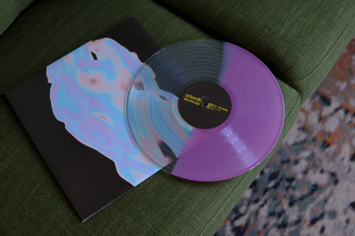 A limited edition Anberlin Silverline clear purple vinyl sits partially on its album cover, which has an abstract pastel face. Both are placed on a green fabric couch next to a patterned rug.