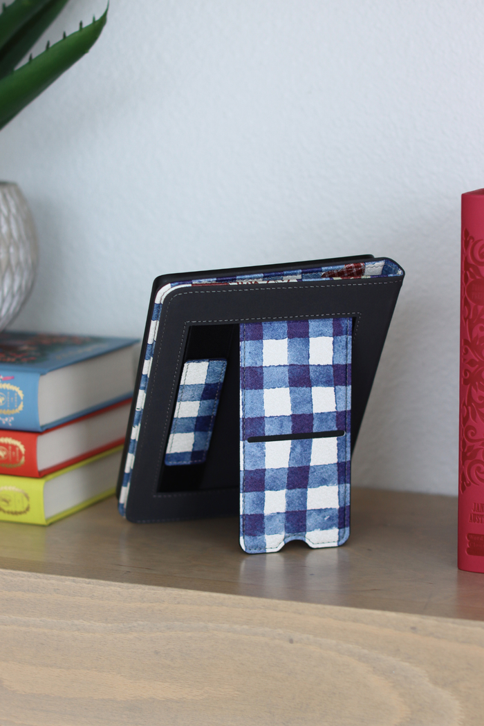 Blue and white checkered Kindle on a wooden surface with books and a plant in the background