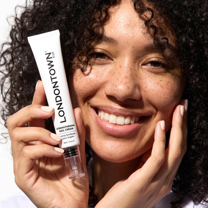 A smiling person with curly hair holds a tube of LONDONTOWN Strengthening Nail Cream close to their face, showcasing the nourishing product that strengthens nails and delivers visible results.