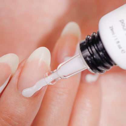A dropper applies white liquid Strengthening Nail Cream by LONDONTOWN to a fingernail, while several natural, manicured nails can be seen on the hand in the background.