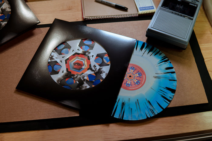 A limited edition Armor For Sleep vinyl with a blue and white splatter design is partially pulled from a black, geometric-art sleeve, resting on a desk next to a retro cassette recorder and stationery.