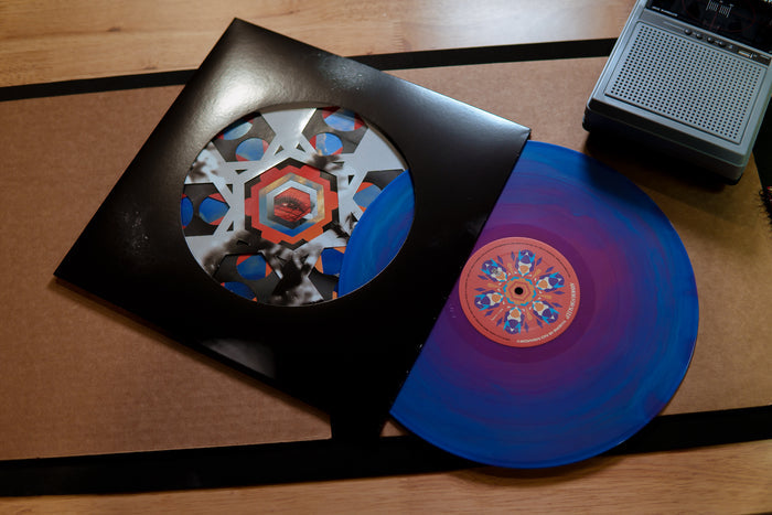 An Armor For Sleep blue and purple vinyl record with a vibrant geometric label is partially pulled from its black die-cut jacket, resting on a wooden table beside a portable tape recorder.