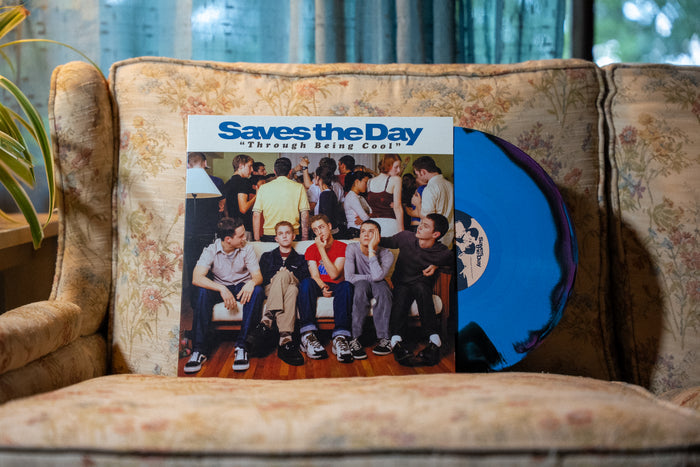 A blue vinyl record and its cover, Saves The Day – Through Being Cool, sit on a floral-patterned couch. The cover features a group of young people at a party.