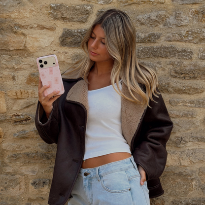 Woman holding a phone up taking a photo, showing a light pale pink phone case with tarot cards and tiny stars. The woman is wearing a brown aviator jacket and light blue jeans.