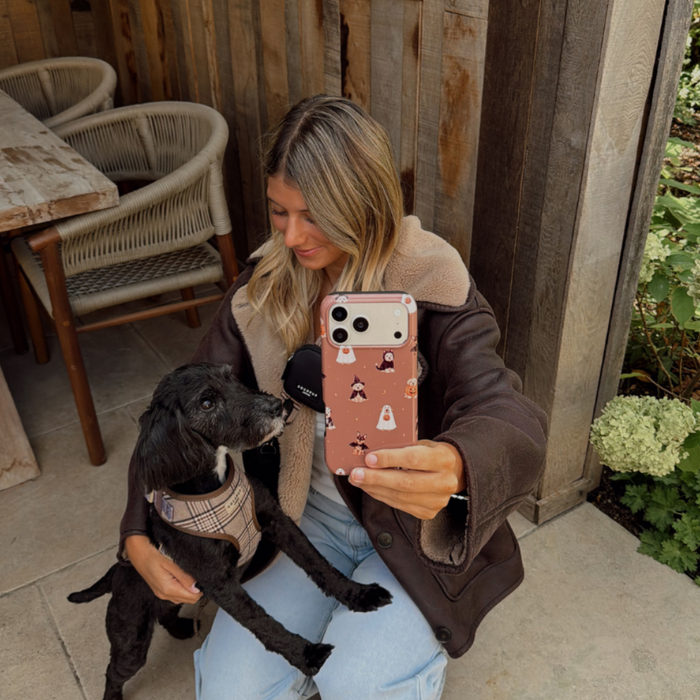 Woman in a brown aviator jacket, holding a phone case with dogs wearing spooky outfits on a brown background, whilst kneeling on the ground stroking a black dog.