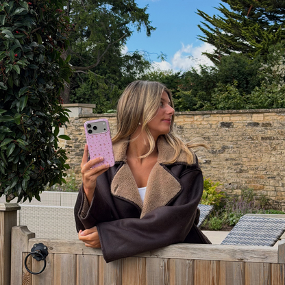 Woman wearing brown aviator jacket holds up an iphone , whilst leaning over a wooden gate, showing a baby pink phone case, with small dainty pastries, pumpkins, wooly hats, bows and leaves printed across the case.