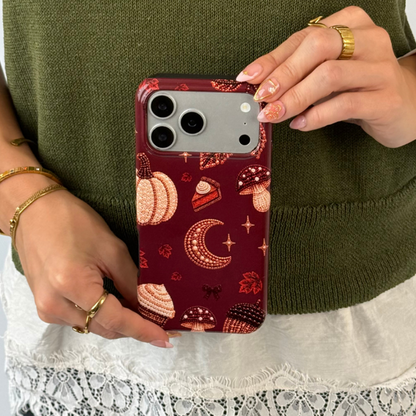 A person holding a phone with a dark red autumn-themed case featuring illustrations of pumpkins, a crescent moon, and a slice of pumpkin pie.