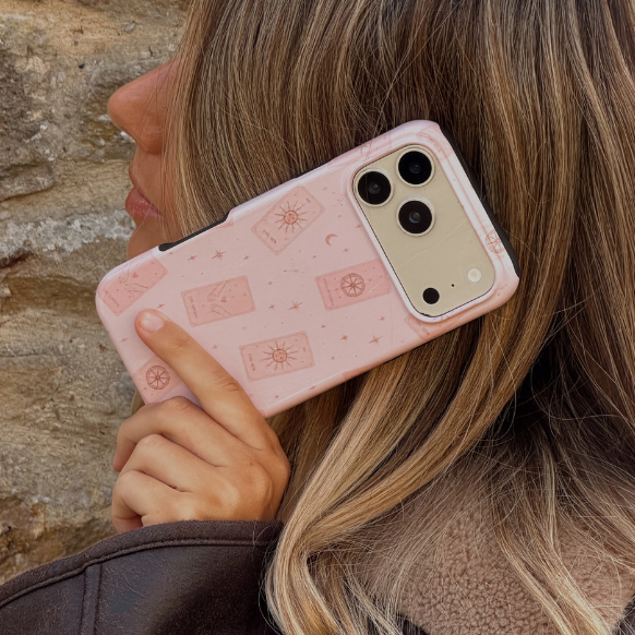 Woman holding a phone up to her ear, showing a light pale pink phone case with tarot cards and tiny stars. The woman is wearing a brown aviator jacket.